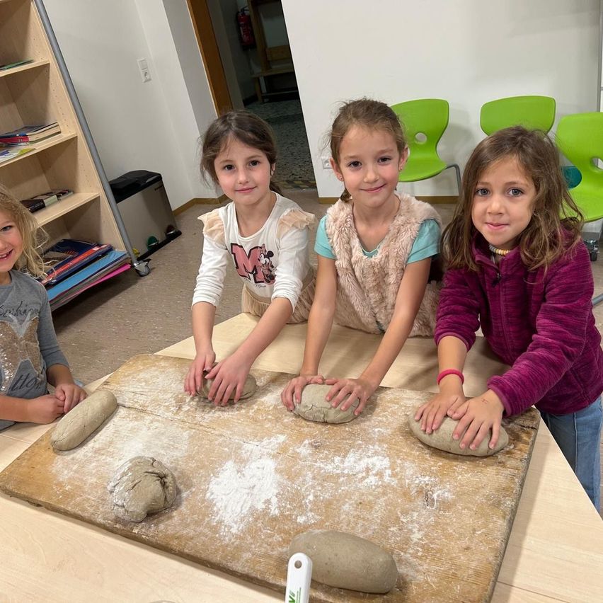 Vier junge Mädchen sitzen an einem Tisch, lächeln und posieren für ein Foto. Sie machen Teig auf einem Holzschneidebrett.