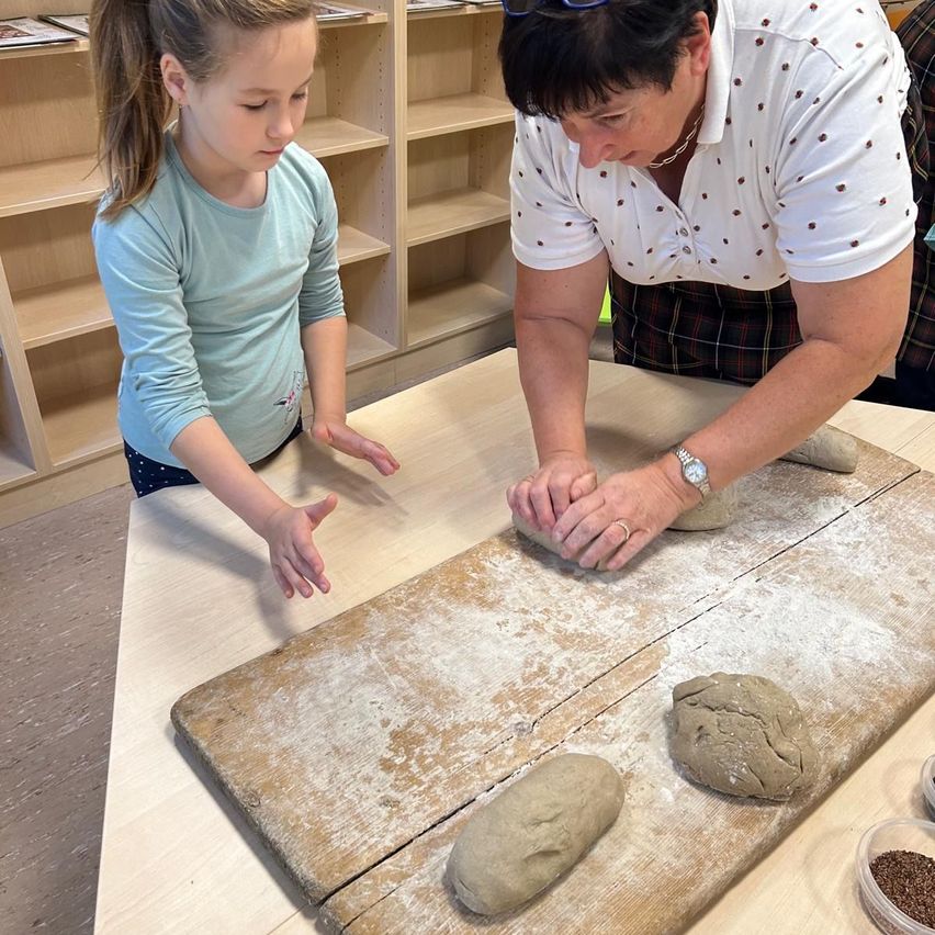 Eine Frau hilft einem jungen Mädchen, Teig auf einem Holzbrett zu formen, Mehl bedeckt den Tisch. Sie sind in einem Raum mit Regalen im Hintergrund.