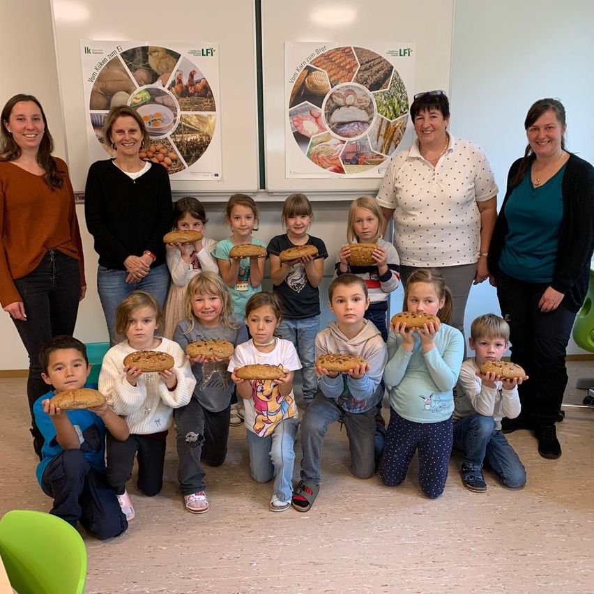 Eine Gruppe von Kindern und Erwachsenen posiert für ein Foto im Klassenzimmer und hält Brot. Sie stehen und sitzen in einer Reihe. Drei Erwachsene stehen hinter ihnen. Hinter ihnen hängen zwei Poster an der Wand.