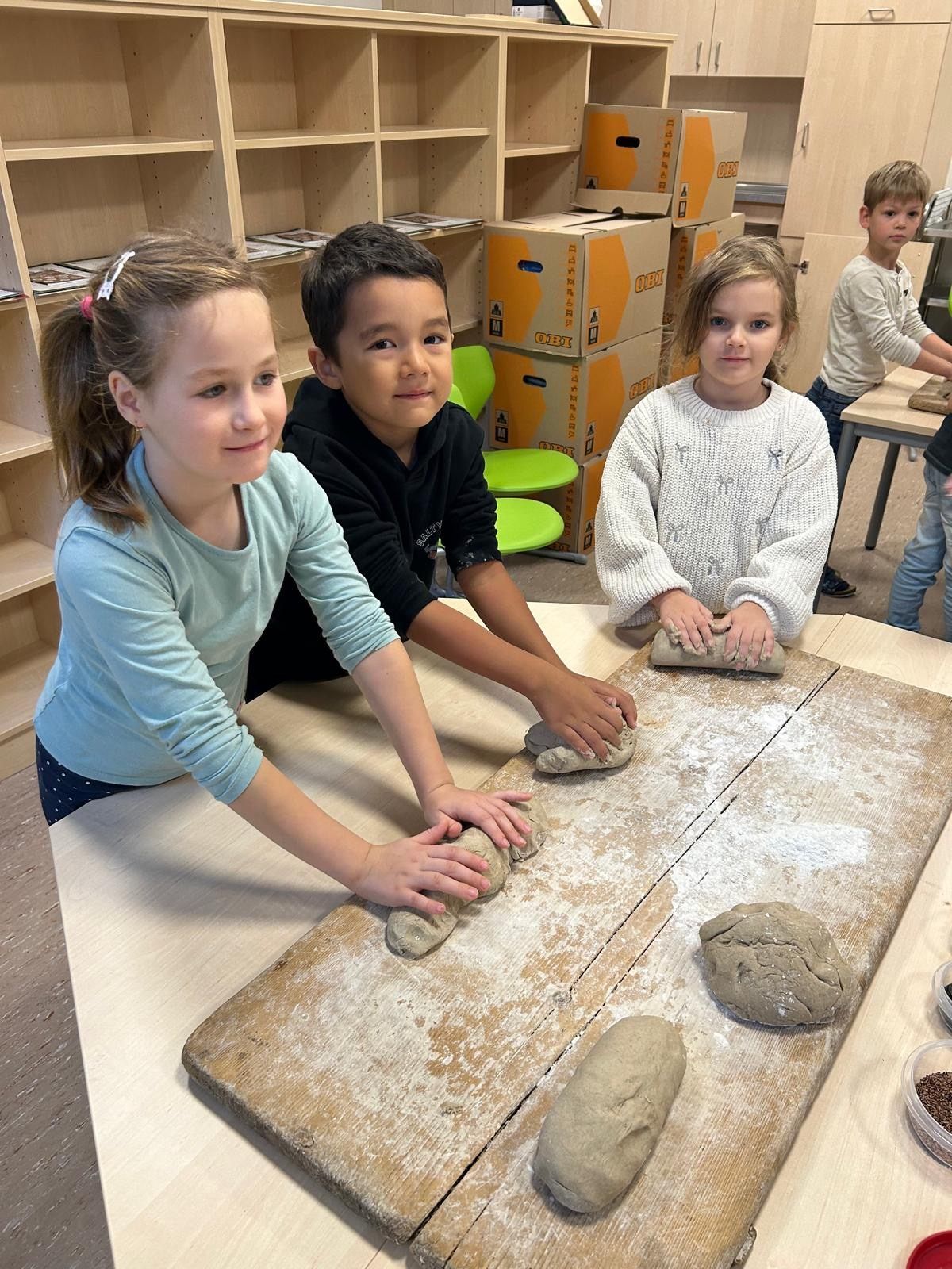 Drei Kinder, zwei Mädchen und ein Junge, sind in einem Klassenzimmer und machen Teig. Das Mädchen auf der linken Seite trägt ein blaues Hemd, der Junge in der Mitte trägt ein schwarzes Hemd und das Mädchen auf der rechten Seite trägt eine weiße Strickjacke. Sie haben mehrere Teigstücke auf einem Holzbrett.