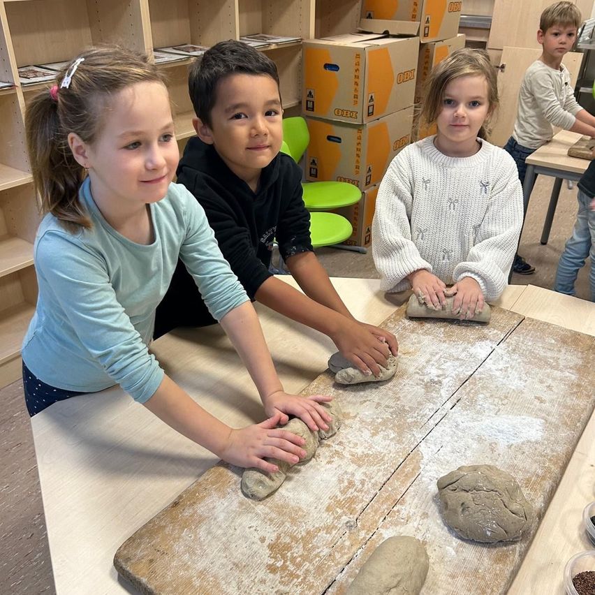 Drei Kinder, zwei Mädchen und ein Junge, sind in einem Klassenzimmer und machen Teig. Das Mädchen auf der linken Seite trägt ein blaues Hemd, der Junge in der Mitte trägt ein schwarzes Hemd und das Mädchen auf der rechten Seite trägt eine weiße Strickjacke. Sie haben mehrere Teigstücke auf einem Holzbrett.