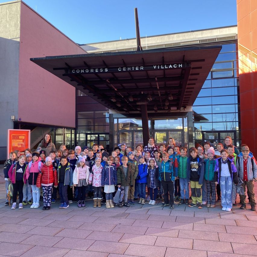 Eine große Gruppe von Kindern und zwei Erwachsenen posiert für ein Foto vor dem Congress Center Villach.