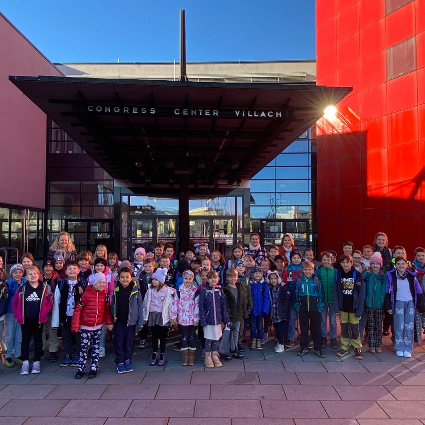 Eine Gruppe von Kindern und Erwachsenen steht vor einem roten Gebäude mit der Aufschrift Congress Center Villach, mit einem glänzenden Eingang.