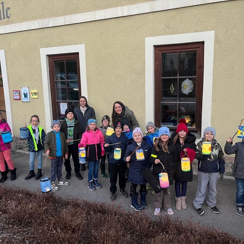 Eine Gruppe von Kindern und Erwachsenen steht vor einem Gebäude, hält Laternen, lächelt und posiert für ein Foto.