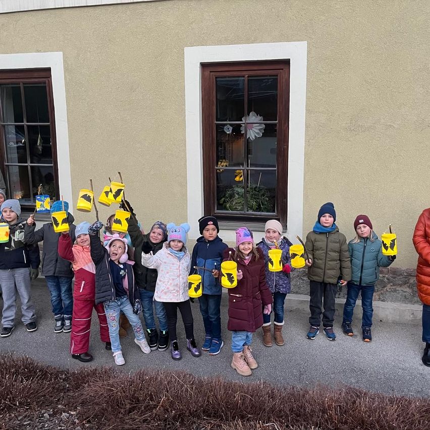 Eine Gruppe von Kindern in Winterkleidung hält gelbe Eimer, wahrscheinlich zum Sammeln von etwas, vor einem Gebäude mit Fenster und Tür.