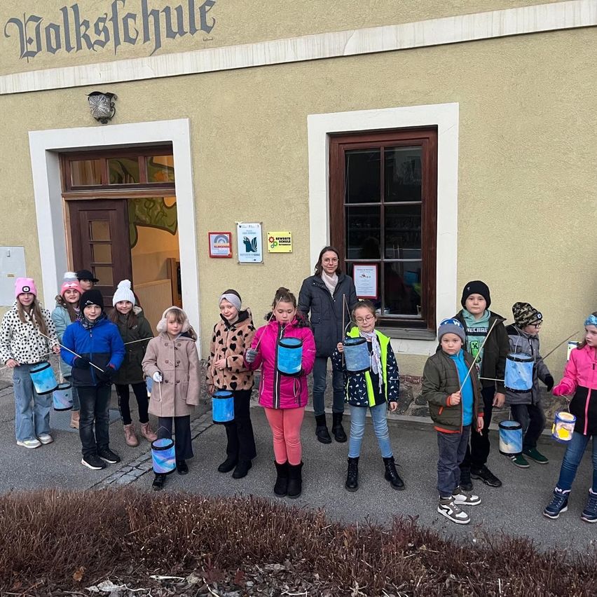Eine Gruppe von Kindern in Winterkleidung hält blaue Eimer und steht vor einem Gebäude mit der Aufschrift 'Volksschule' oben.