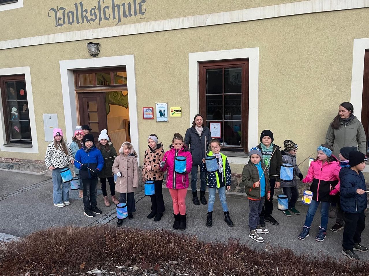 Eine Gruppe von Kindern in Winterkleidung hält blaue Eimer und steht vor einem Gebäude mit der Aufschrift 'Volksschule' oben.