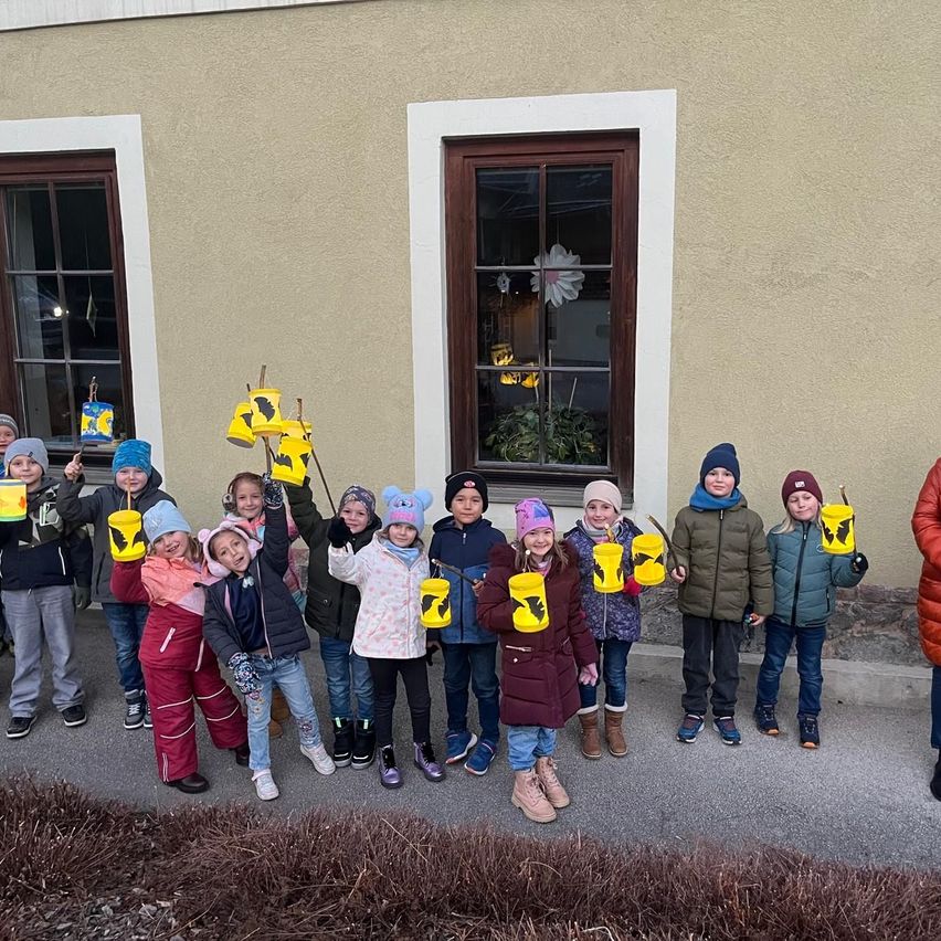 Eine Gruppe von Kindern in Winterkleidung, die gelbe Eimer halten, steht vor einem Gebäude mit zwei Fenstern.