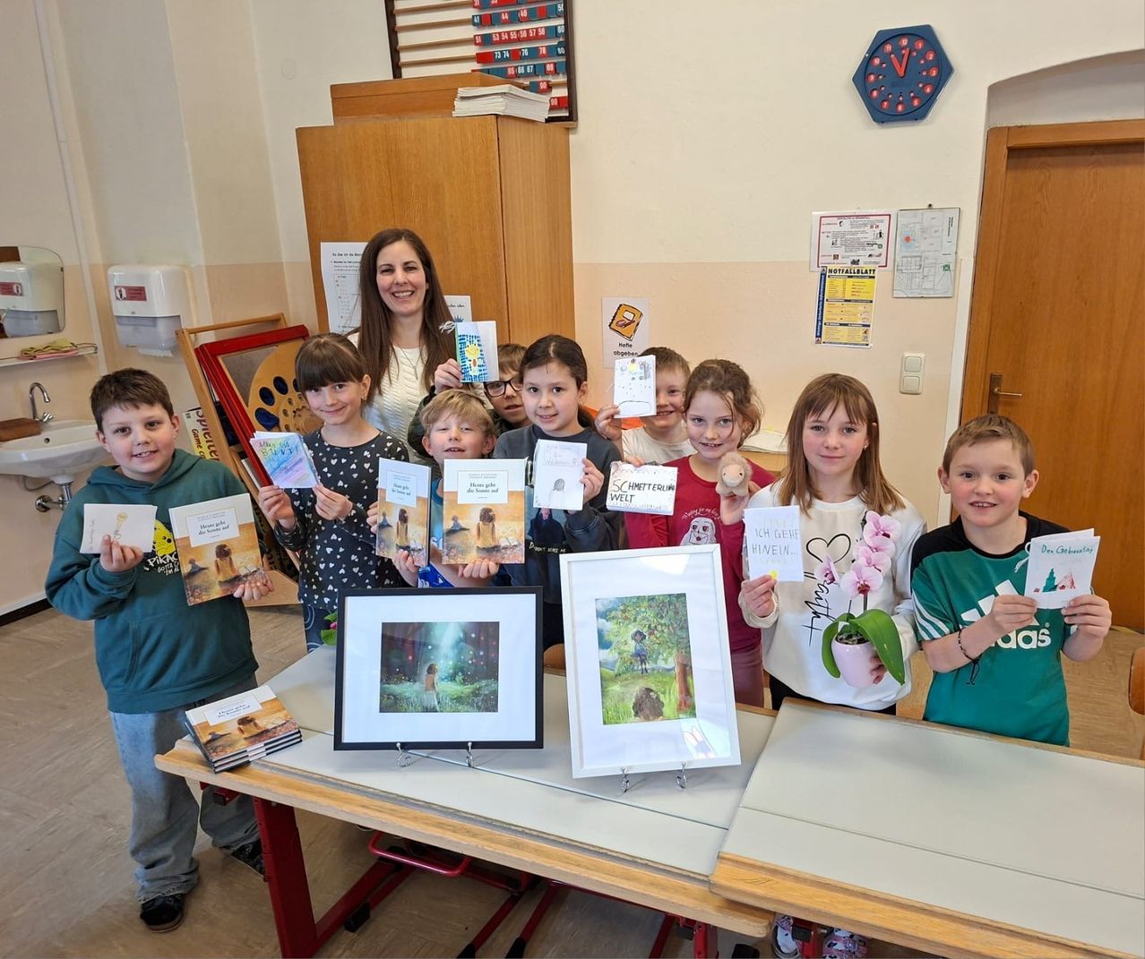 Eine Gruppe von Kindern und einer Frau stehen in einem Klassenzimmer, halten Bücher und gerahmte Bilder und lächeln für ein Foto. Hinter ihnen hängen eine Uhr und verschiedene Poster an der Wand.