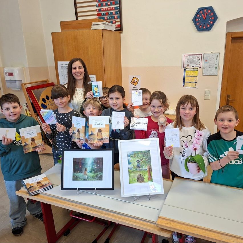 Eine Gruppe von Kindern und einer Frau stehen in einem Klassenzimmer, halten Bücher und gerahmte Bilder und lächeln für ein Foto. Hinter ihnen hängen eine Uhr und verschiedene Poster an der Wand.