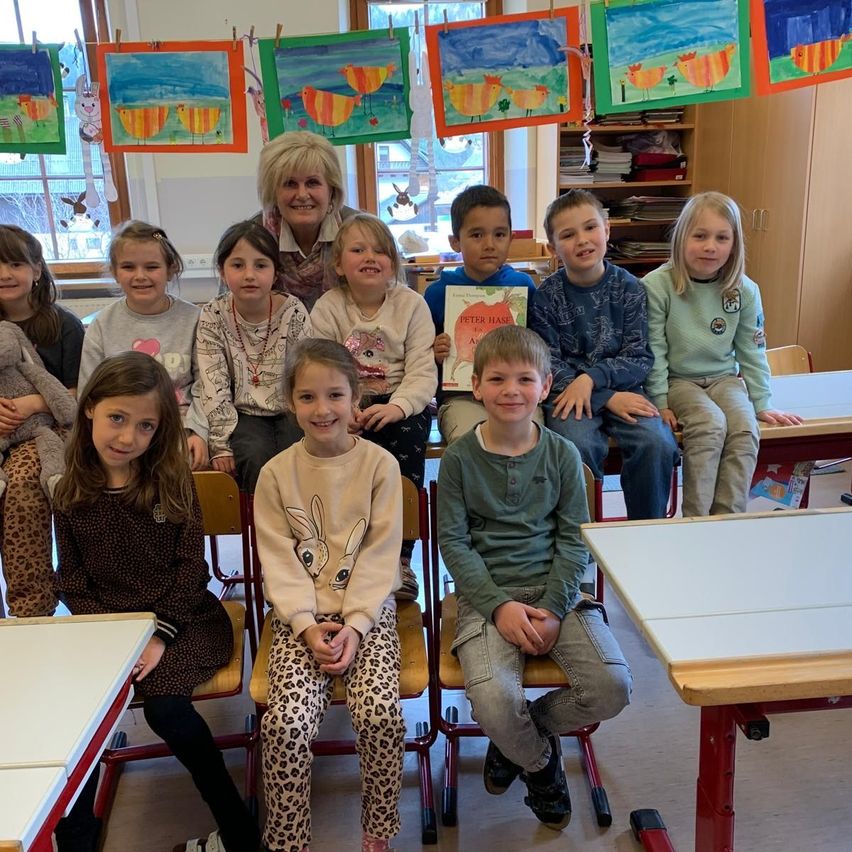 Eine Gruppe von Kindern und ihrer Lehrerin in einem Klassenzimmer mit farbenfrohen Kunstwerken an der Wand. Sie sitzen auf Stühlen und lächeln für ein Foto.