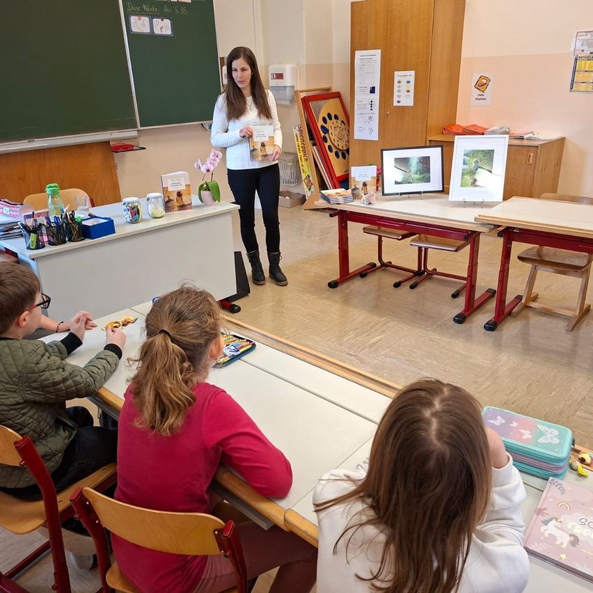 Eine Frau steht in einem Klassenzimmer, hält ein Buch in den Händen, während vier Kinder an Tischen sitzen. Der Raum ist mit Holzmöbeln ausgestattet und zeigt verschiedene Gegenstände an der Wand.