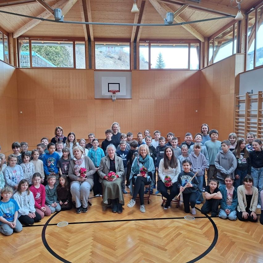 Gruppenfoto von Schülern und Lehrern in einer Turnhalle mit einem Basketballkorb an der Wand und einer Frau mit Blumen in der Mitte sitzend.