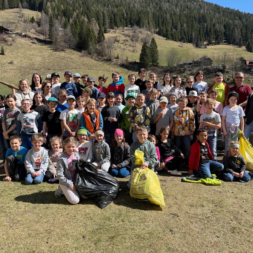 Eine große Gruppe von Kindern und Erwachsenen posiert für ein Foto auf einer Grasfläche. Sie haben Müllsäcke und tragen Freizeitkleidung. Der Hintergrund zeigt einen bewaldeten Hügel.