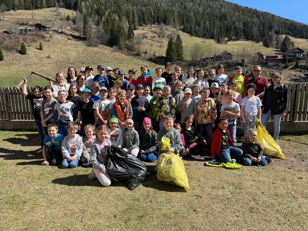 Eine große Gruppe von Kindern und Erwachsenen posiert für ein Foto auf einer Grasfläche. Sie haben Müllsäcke und tragen Freizeitkleidung. Der Hintergrund zeigt einen bewaldeten Hügel.
