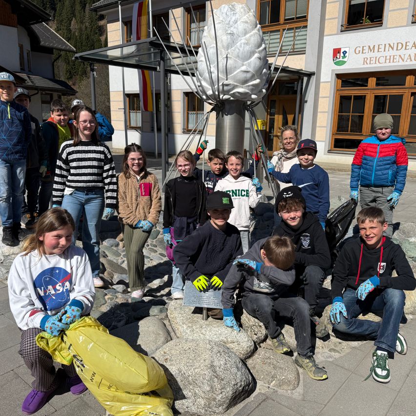 Eine Gruppe von Schülern und Erwachsenen versammelt sich um eine Skulptur vor einem Gebäude, trägt Handschuhe und hält Plastiktüten in der Hand. Sie alle lächeln und posieren für ein Foto.