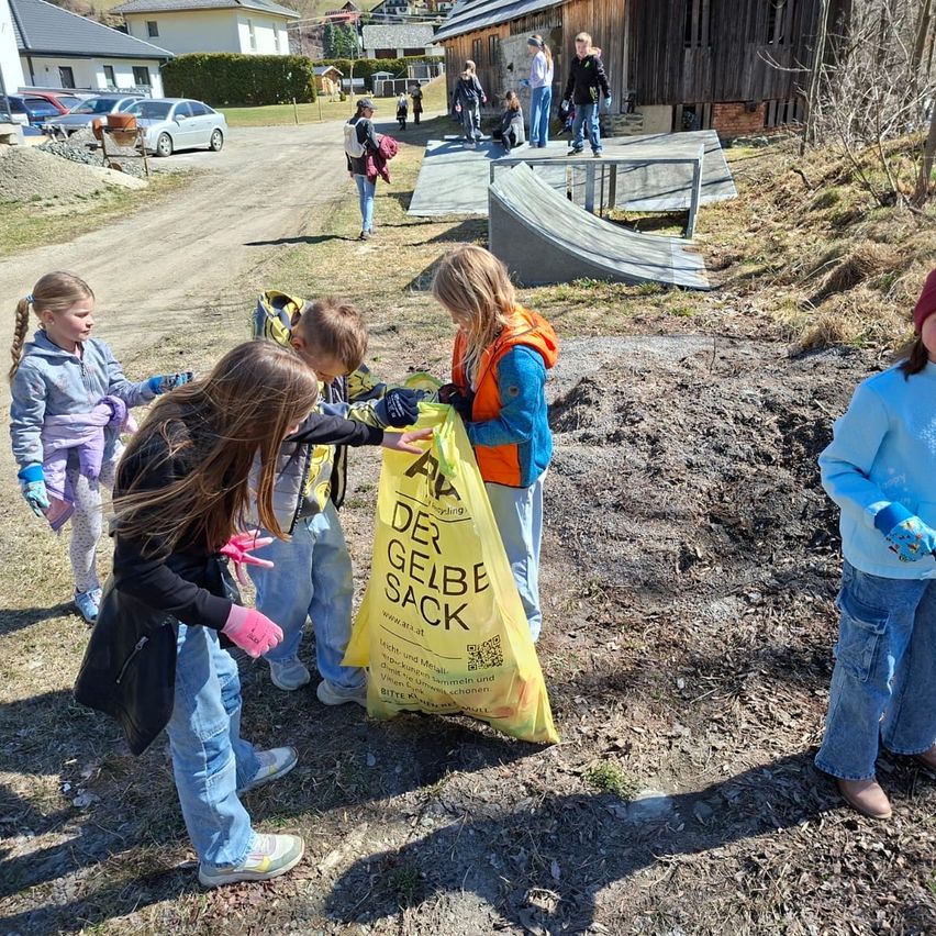 Eine Gruppe von Kindern sammelt Müll in einem staubigen Bereich und trägt Handschuhe. Ein gelber Beutel mit der Aufschrift 'Der gelbe Sack' ist im Vordergrund. Einige Leute laufen auf der Straße.
