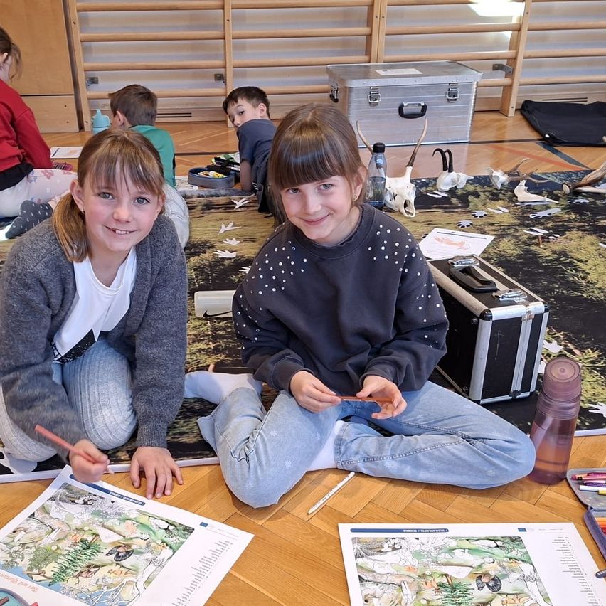 Zwei junge Mädchen sitzen auf dem Boden und beschäftigen sich mit dem Ausmalen eines Bildes. Eine benutzt einen Bleistift und die andere einen Marker. Sie sind von Malbüchern und Materialien umgeben. Hinter ihnen malen auch andere Kinder.