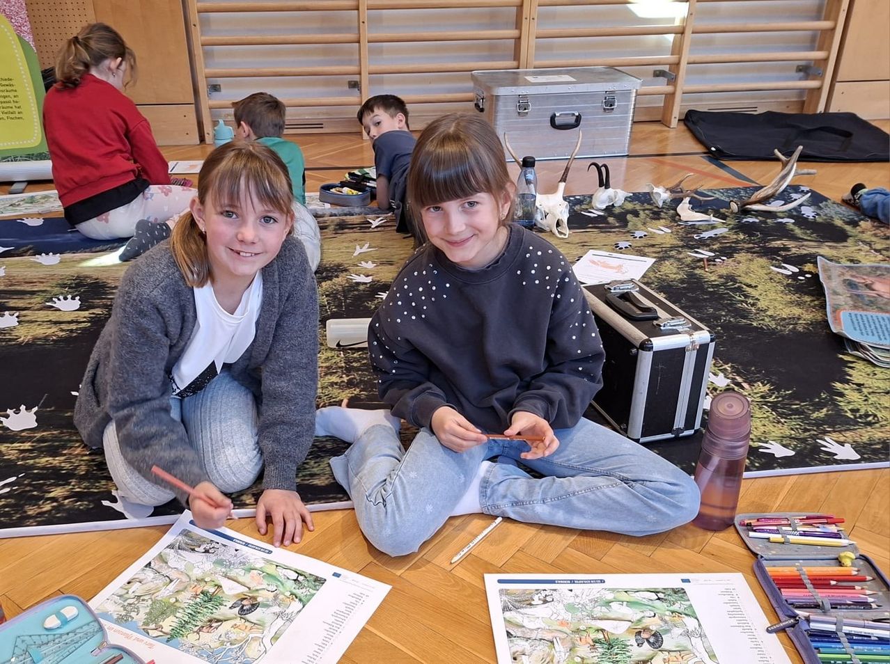 Zwei junge Mädchen sitzen auf dem Boden und beschäftigen sich mit dem Ausmalen eines Bildes. Eine benutzt einen Bleistift und die andere einen Marker. Sie sind von Malbüchern und Materialien umgeben. Hinter ihnen malen auch andere Kinder.