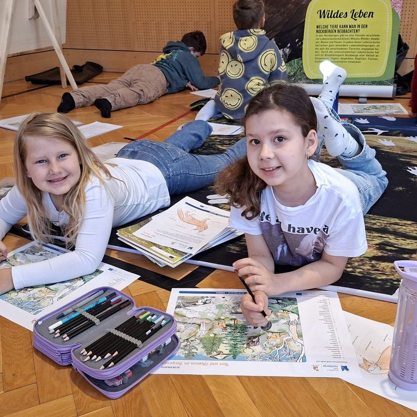 Zwei Mädchen sitzen lächelnd auf dem Boden und machen eine Malaktivität. Das Mädchen auf der rechten Seite hält einen Stift und malt ein Buch aus. Das Mädchen auf der linken Seite hält einen Stiftekasten. Es gibt einige Papiere, Stifte und andere Gegenstände auf dem Boden. Hinter ihnen liegt ein Junge auf dem Boden.