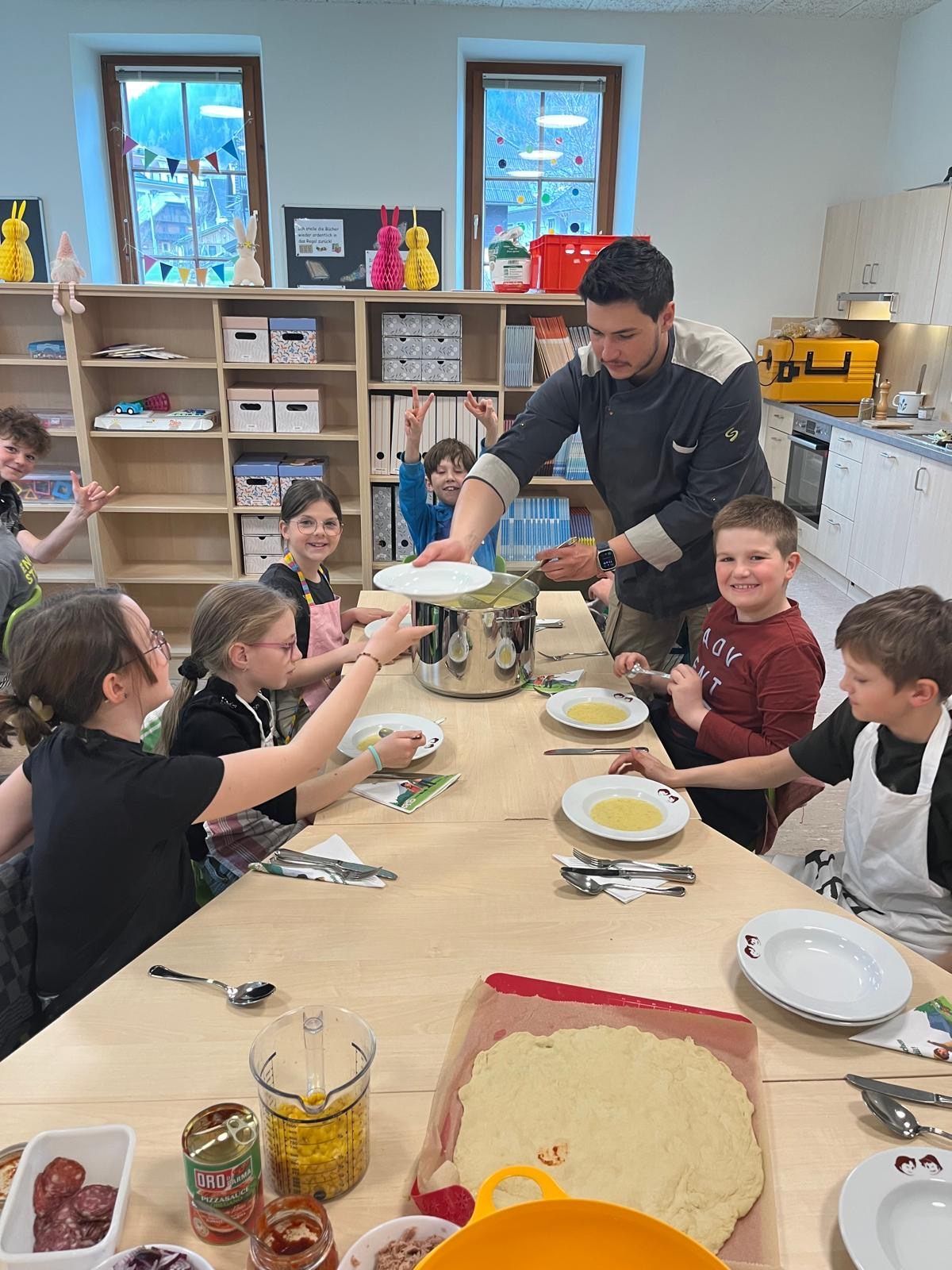 Ein Koch steht über einem Tisch, an dem Kinder sitzen, die eine Schüssel halten, während sie scheinbar Essen zubereiten. Einige Kinder tragen Schürzen und haben Teller vor sich.