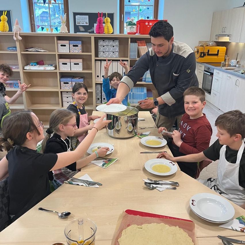 Ein Koch steht über einem Tisch, an dem Kinder sitzen, die eine Schüssel halten, während sie scheinbar Essen zubereiten. Einige Kinder tragen Schürzen und haben Teller vor sich.