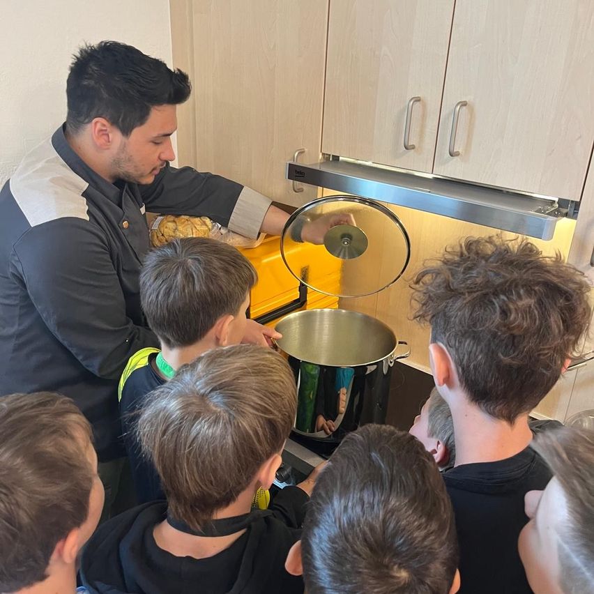Ein Mann in einer Küche mit mehreren Jungen um ihn herum. Er hält einen Topfdeckel in der Hand und bereitet das Kochen vor. Die Kinder beobachten aufmerksam.