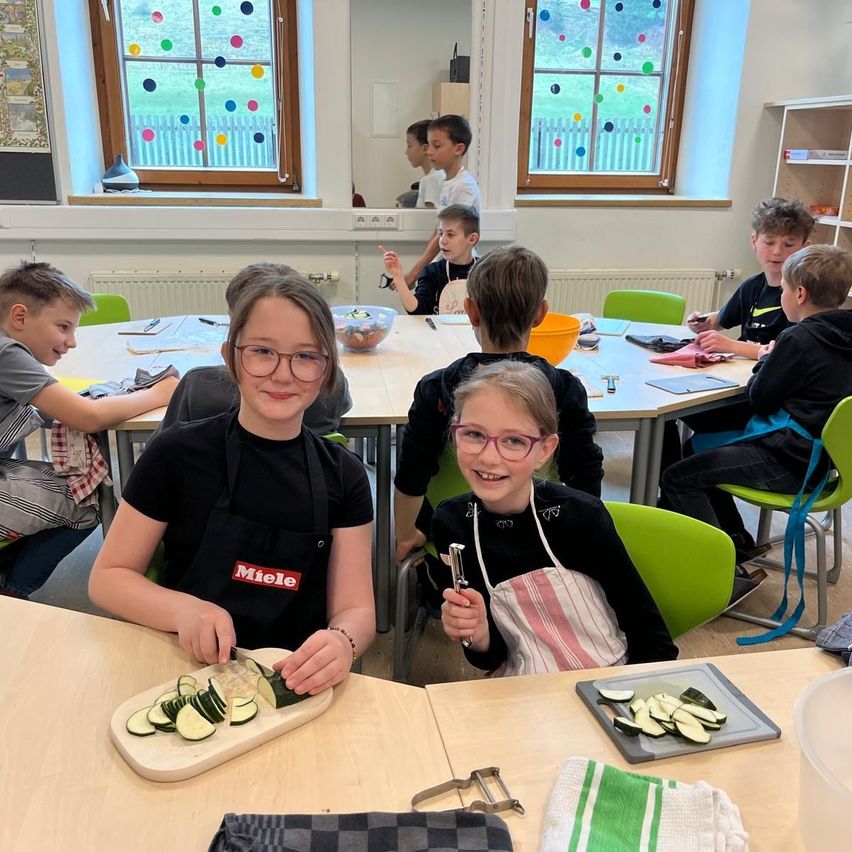 Kinder in einem Klassenzimmer sind an einer Kochaktivität beteiligt. Zwei Mädchen schneiden Gurken auf einem Schneidebrett, wobei eines ein Miele-Schürze trägt. Andere Kinder sitzen um einen Tisch herum, einige mit Schürzen, und einer hält eine Schüssel. Das Klassenzimmer ist gut beleuchtet mit farbenfrohen Fenstern.