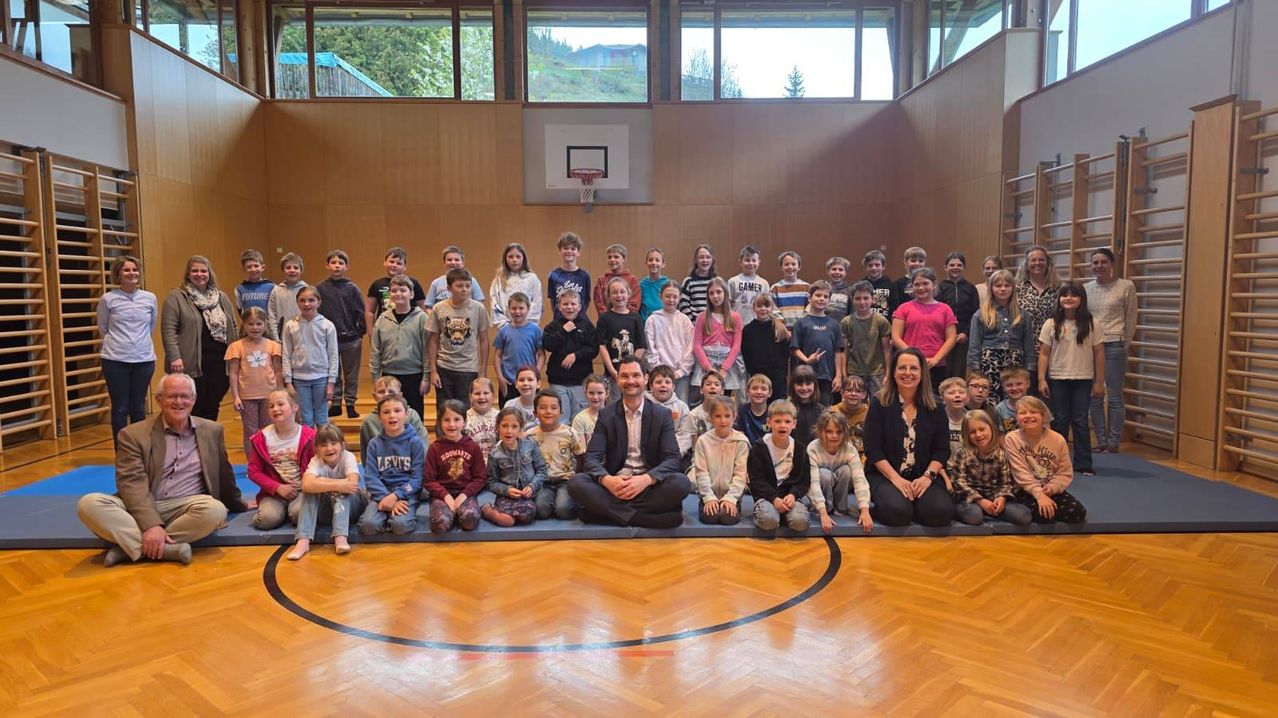 Eine Gruppe von Kindern und Erwachsenen posiert für ein Foto auf einem überdachten Basketballplatz. Die Kinder sitzen auf dem Boden, während ein Mann kniet.
