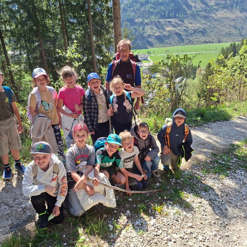 Eine Gruppe von Kindern und einem Erwachsenen posiert für ein Foto auf einem Bergpfad, umgeben von Bäumen und einem Tal in der Ferne.