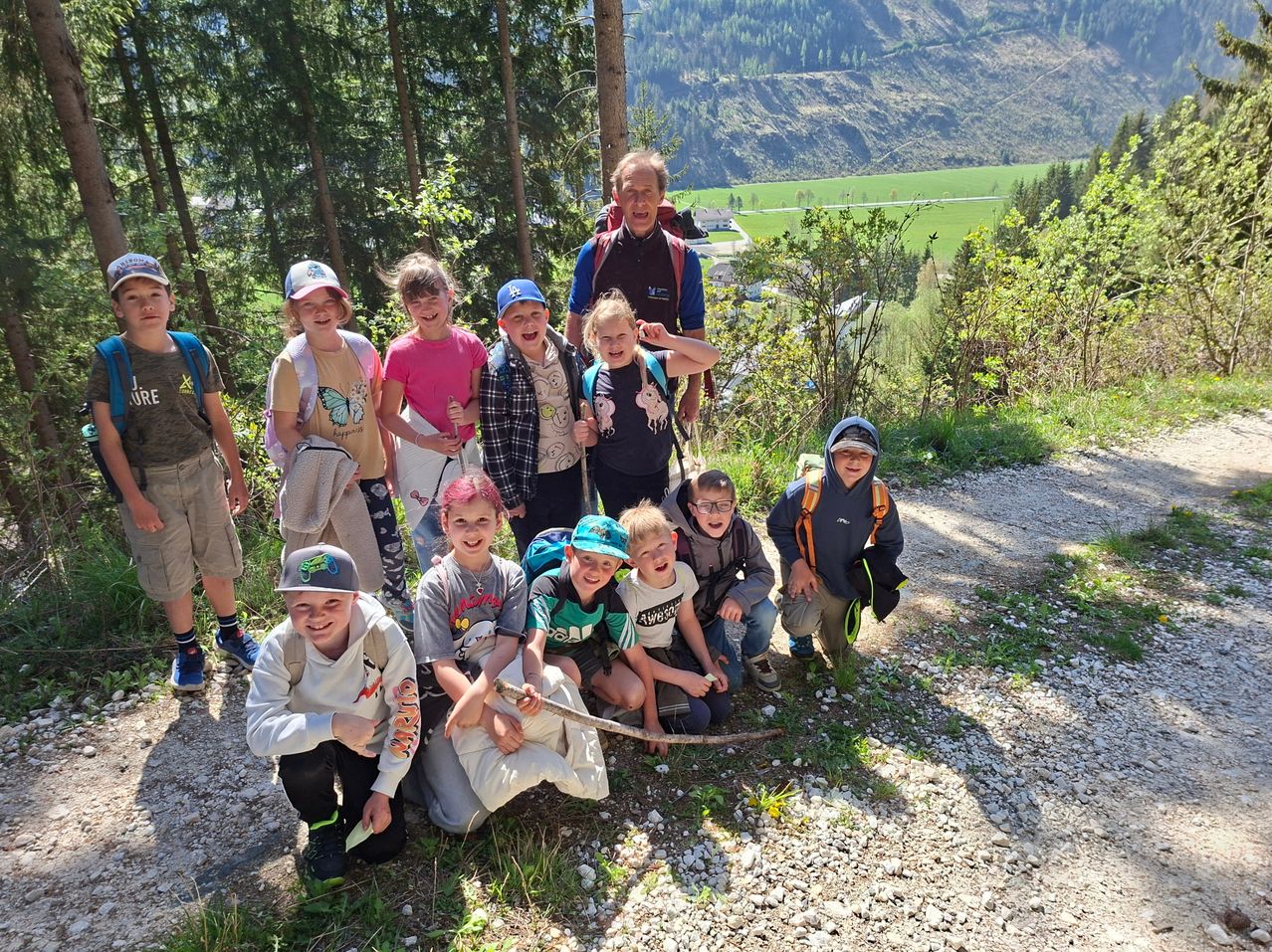 Eine Gruppe von Kindern und einem Erwachsenen posiert für ein Foto auf einem Bergpfad, umgeben von Bäumen und einem Tal in der Ferne.