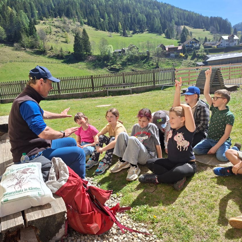 Ein Mann spricht zu einer Gruppe von Kindern, die auf dem Gras in einer ländlichen Gegend sitzen. Einige Kinder haben die Hände erhoben. Ein roter Rucksack mit dem Text 'Naturschutz in Kärnten' liegt in der Nähe.