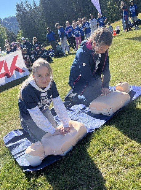 Zwei Frauen üben CPR auf einem Dummies auf einem Rasenfeld, während ein Publikum im Hintergrund zuschaut.