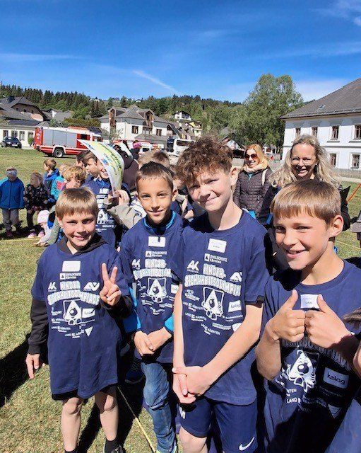 Vier Jungen in blauen Shirts posieren für ein Foto mit erhobenen Daumen an einem sonnigen Tag, mit einer Menschenmenge und Gebäuden im Hintergrund.