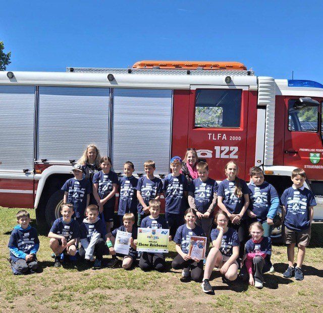 Eine Gruppe von Kindern und Erwachsenen posiert für ein Foto vor einem Feuerwehrwagen. Die Kinder halten Zertifikate in den Händen und einige haben Rucksäcke.