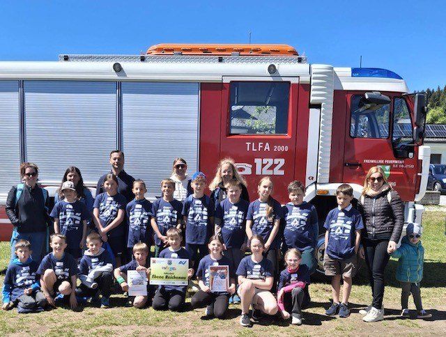 Eine Gruppe von Kindern und Erwachsenen posiert für ein Foto vor einem Feuerwehrfahrzeug mit der Nummer 122. Die Kinder tragen einheitliche blaue T-Shirts, und einige halten Urkunden.