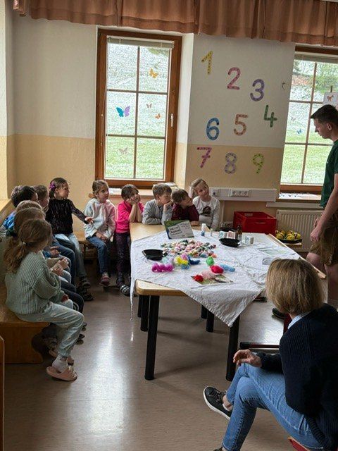 Kinder sitzen um einen Tisch mit Bastelarbeiten, eine Zahlentafel ist an der Wand und zwei Erwachsene sind anwesend.