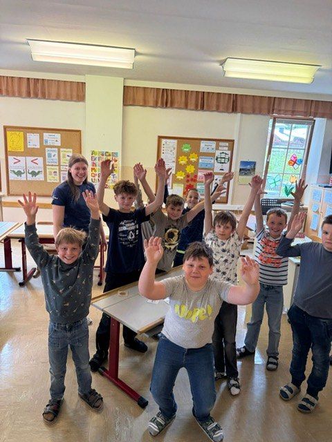 A group of children and a teacher in a classroom, all raising their hands. The teacher stands in front of them. The room has white walls, bulletin boards, and windows.