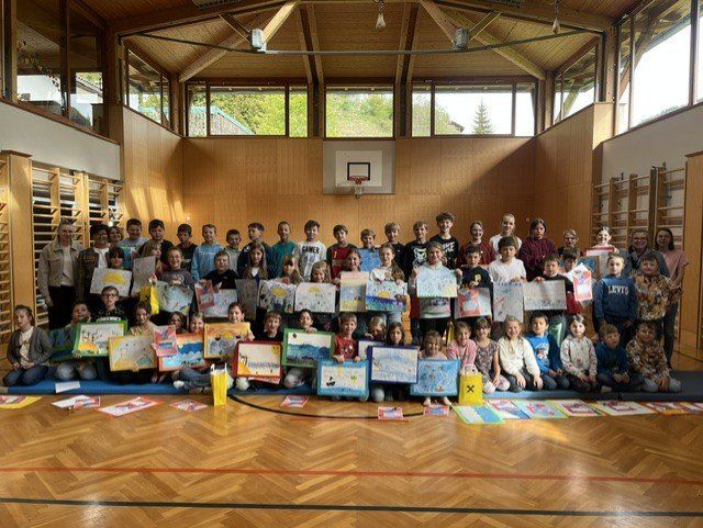 Eine Gruppe von Kindern posiert für ein Foto in einer Turnhalle, sie halten Zeichnungen mit verschiedenen Kunstwerken hoch, im Hintergrund befindet sich ein Basketballkorb.
