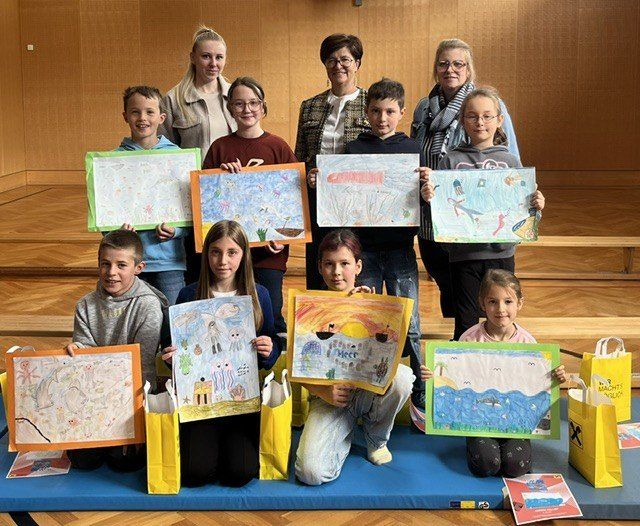 Eine Gruppe von Kindern und Erwachsenen steht und sitzt auf einer Bühne und hält Zeichnungen und Papiertüten in der Hand. Die Zeichnungen zeigen verschiedene Szenen, wie ein Schiff, eine Landschaft und ein Meer mit Fischen. Die Kinder lächeln und posieren für die Kamera.