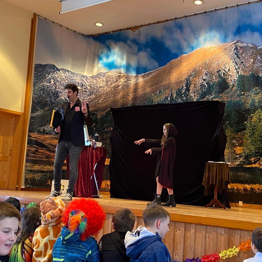Ein Zauberer führt auf der Bühne vor einem Hintergrund von Bergen eine Vorstellung durch. Ein junges Mädchen hilft ihm, und mehrere Kinder beobachten zu.