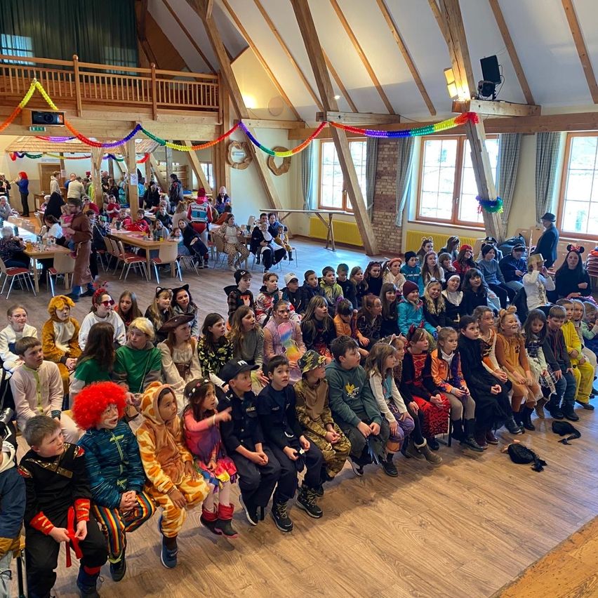 Eine große Gruppe von Kindern in Kostümen versammelt sich in einem Saal. Einige sitzen auf dem Boden, andere stehen. Bunte Dekorationen und eine Bühne sind vorhanden. An der Wand ist ein Lautsprecher montiert.