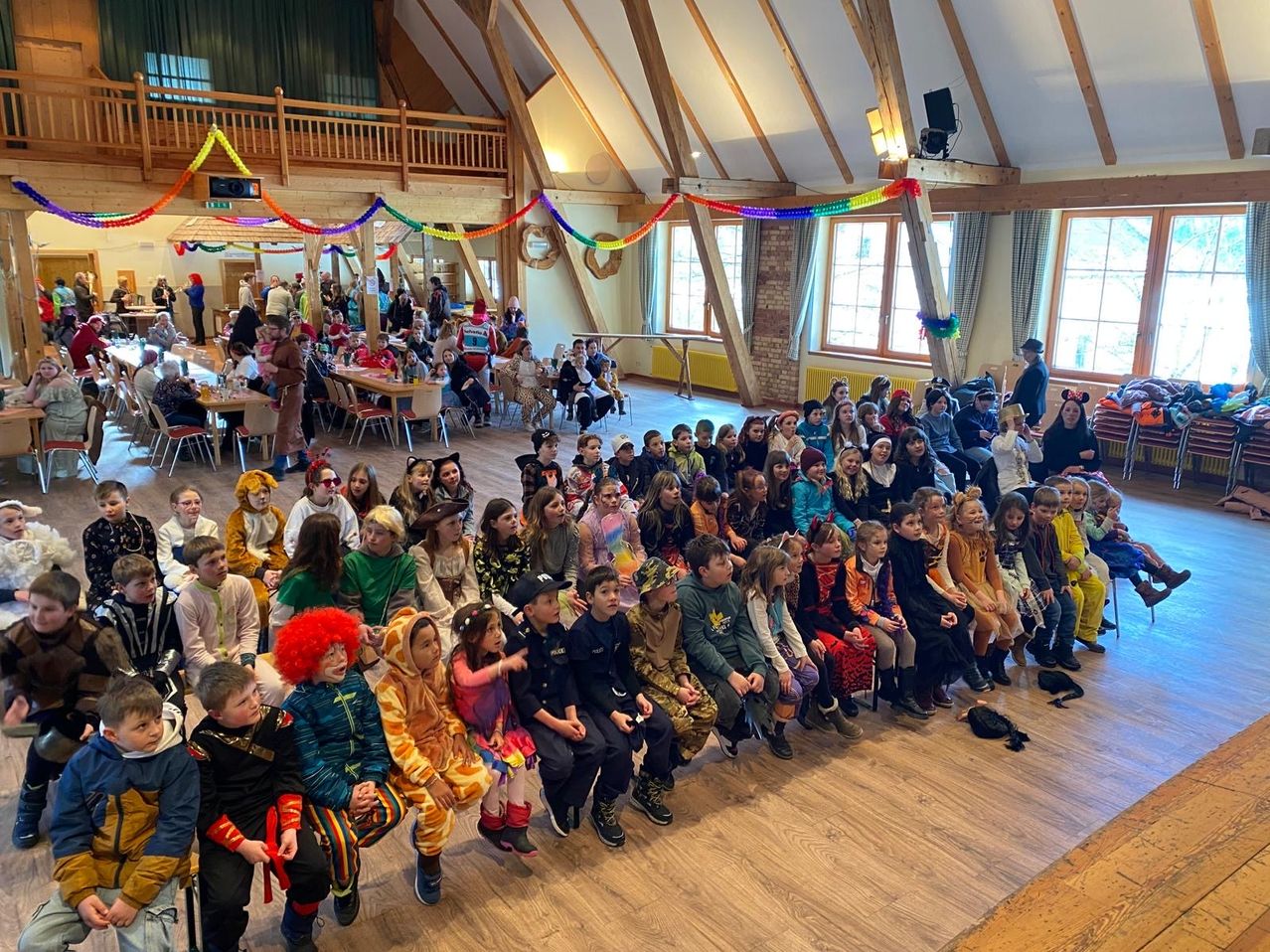 Eine große Gruppe von Kindern in Kostümen versammelt sich in einem Saal. Einige sitzen auf dem Boden, andere stehen. Bunte Dekorationen und eine Bühne sind vorhanden. An der Wand ist ein Lautsprecher montiert.