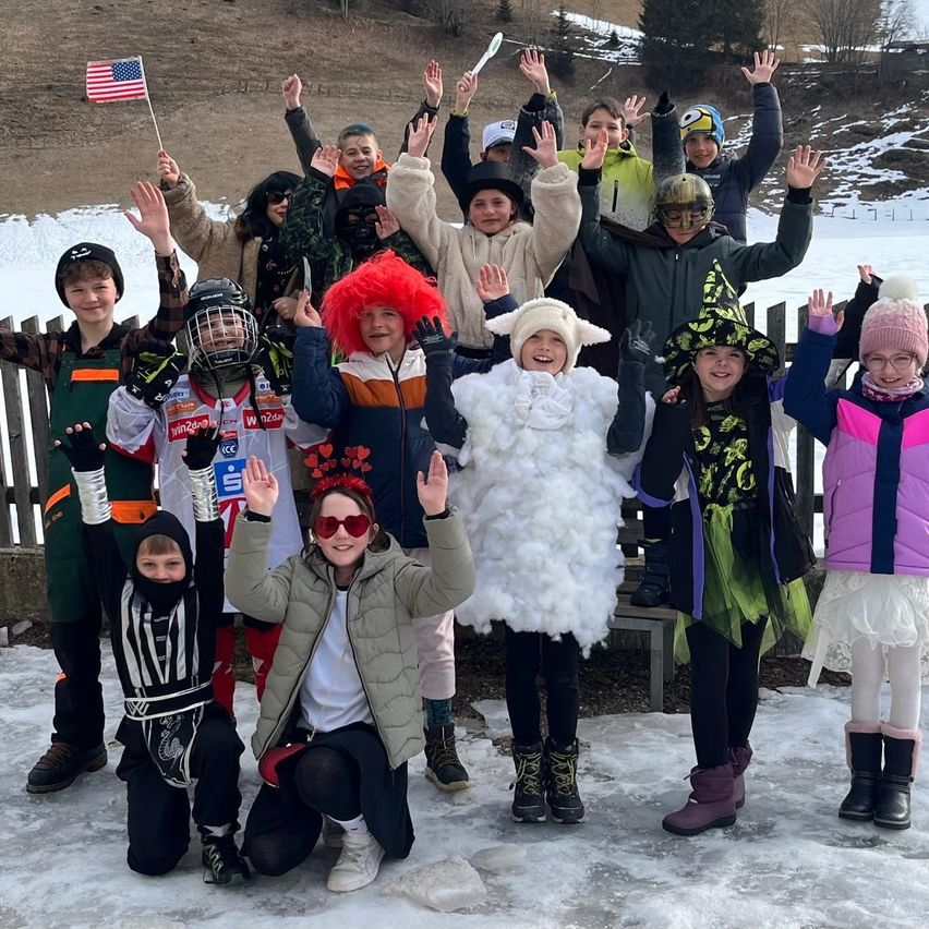 Eine Gruppe von Kindern in verschiedenen Kostümen posiert für ein Foto im Schnee mit einem Holzzaun dahinter. Ein Kind ist als Hexe verkleidet.