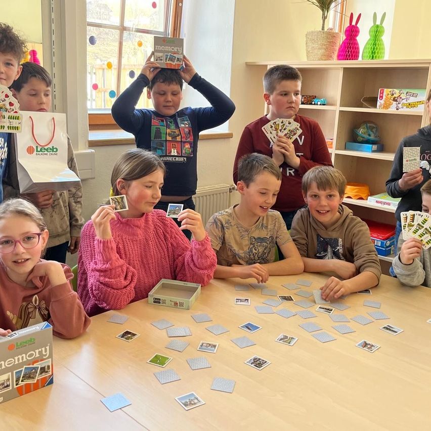 Eine Gruppe von Kindern spielt ein Gedächtniskartenspiel mit Karten auf einem Tisch in einem Raum. Sie lächeln und halten Karten hoch.