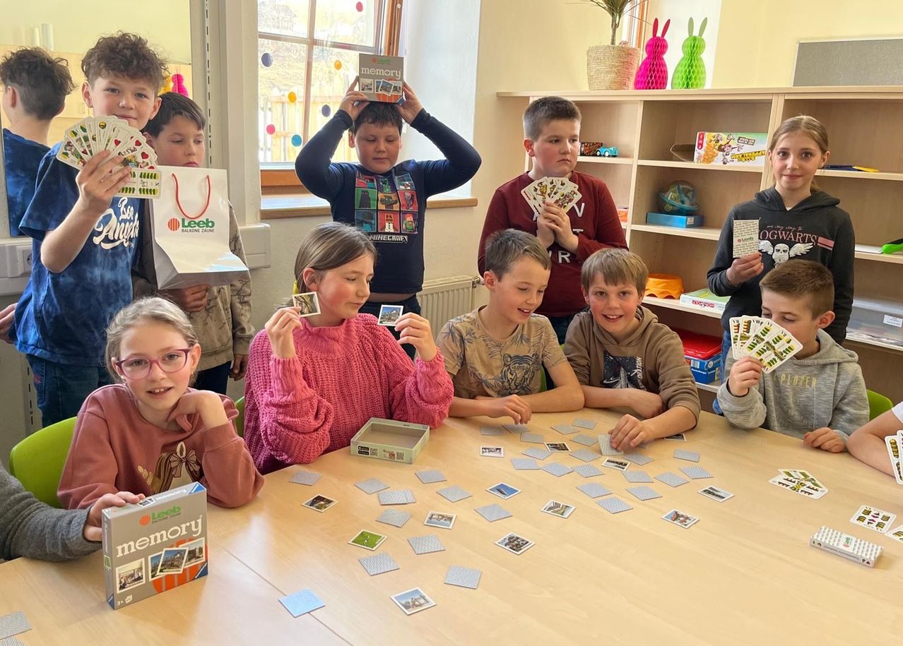 Eine Gruppe von Kindern spielt ein Gedächtniskartenspiel mit Karten auf einem Tisch in einem Raum. Sie lächeln und halten Karten hoch.