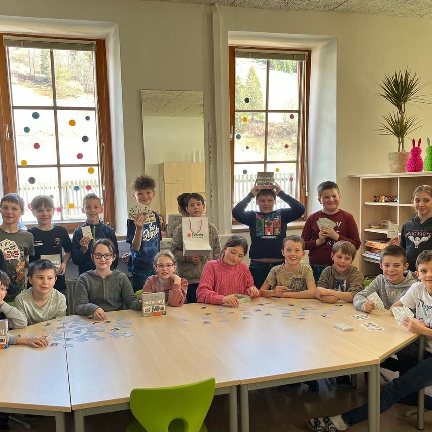 Eine Gruppe von Kindern sitzt um einen Tisch herum und hält Karten in den Händen, einige stehen und lächeln. Ein Junge hält ein Buch über seinem Kopf. Der Raum hat große Fenster mit bunten Mustern und Regale mit Büchern und Pflanzen.