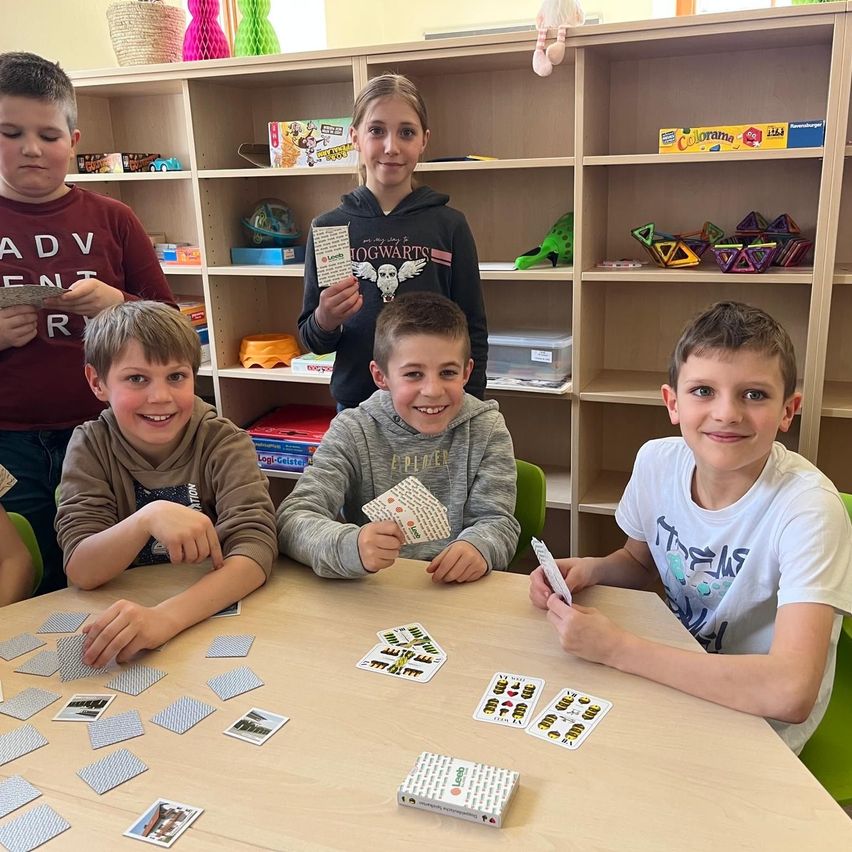 Fünf Kinder sitzen um einen Tisch herum und spielen ein Kartenspiel. Sie lächeln und halten Karten in den Händen. Ein Mädchen in einer Kapuzenjacke steht hinter ihnen. Der Tisch ist mit Karten übersät. Im Hintergrund befinden sich Regale mit verschiedenen Gegenständen, darunter Bücher und Spielzeug.