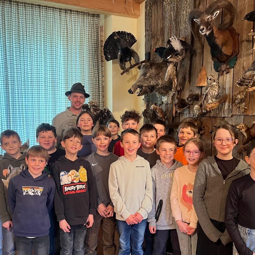 Eine Gruppe von Kindern und zwei Erwachsenen steht vor einer Wand, die mit verschiedenen ausgestopften Tieren verziert ist. Die Kinder lächeln und die Erwachsenen stehen hinten.
