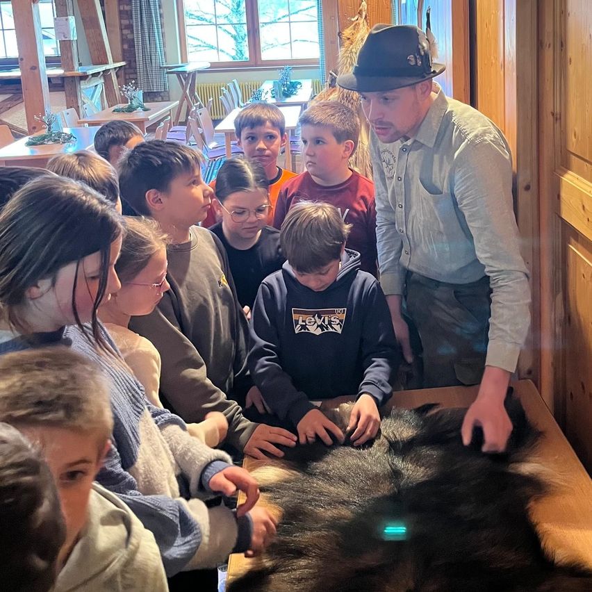 Ein Mann mit Hut unterrichtet Kinder über ein Fell auf einem Tisch in einem Raum mit Holzwänden und Tischen.
